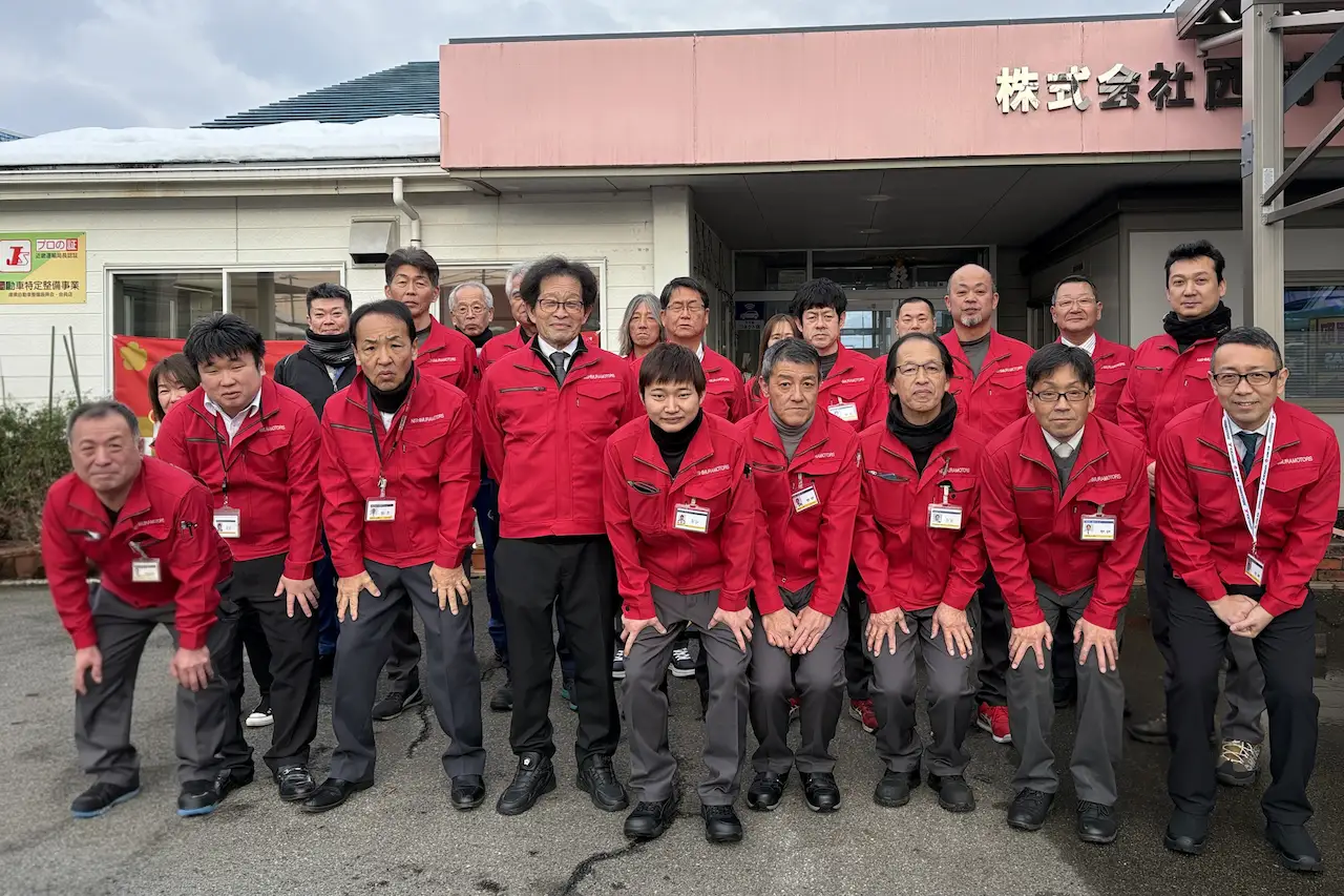 事務所の前で、赤い作業ジャンパーを着用したスタッフ一同が笑顔で並ぶ集合写真