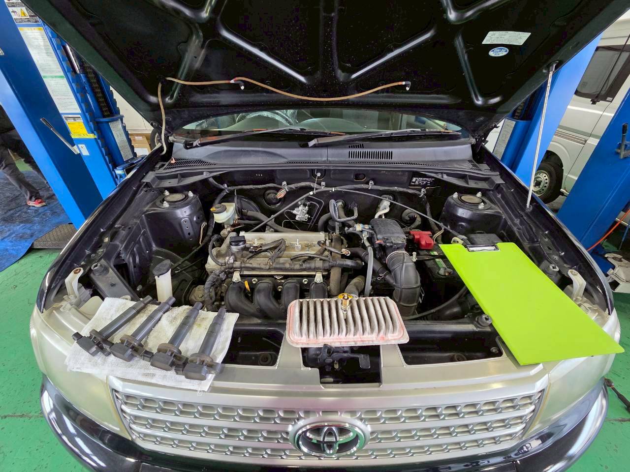 ボンネットが開いた車のエンジンルームと、その手前に取り外されて並べられたエアフィルターやスパークプラグなどの部品